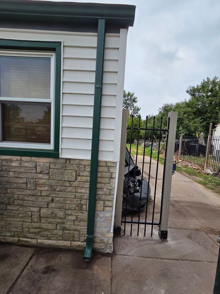 Side yard gate beside a white-and-stone house, with a narrow path and trash bags beyond the fence.