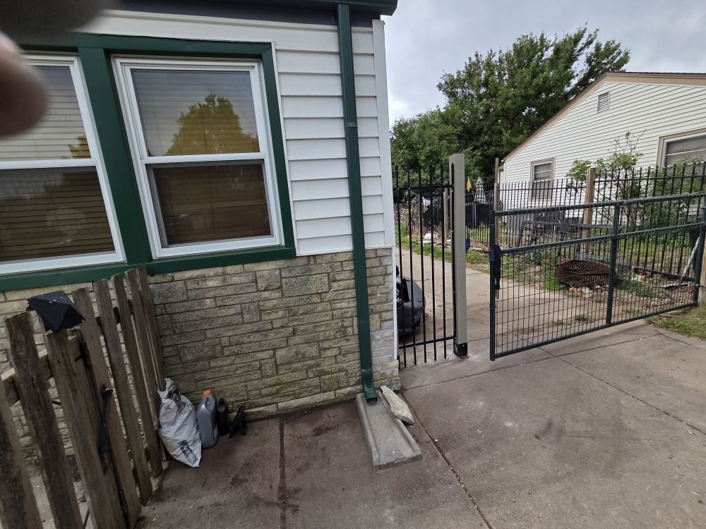 House side yard with stone-trimmed wall, wooden fence, and open black metal gate leading to a concrete patio.