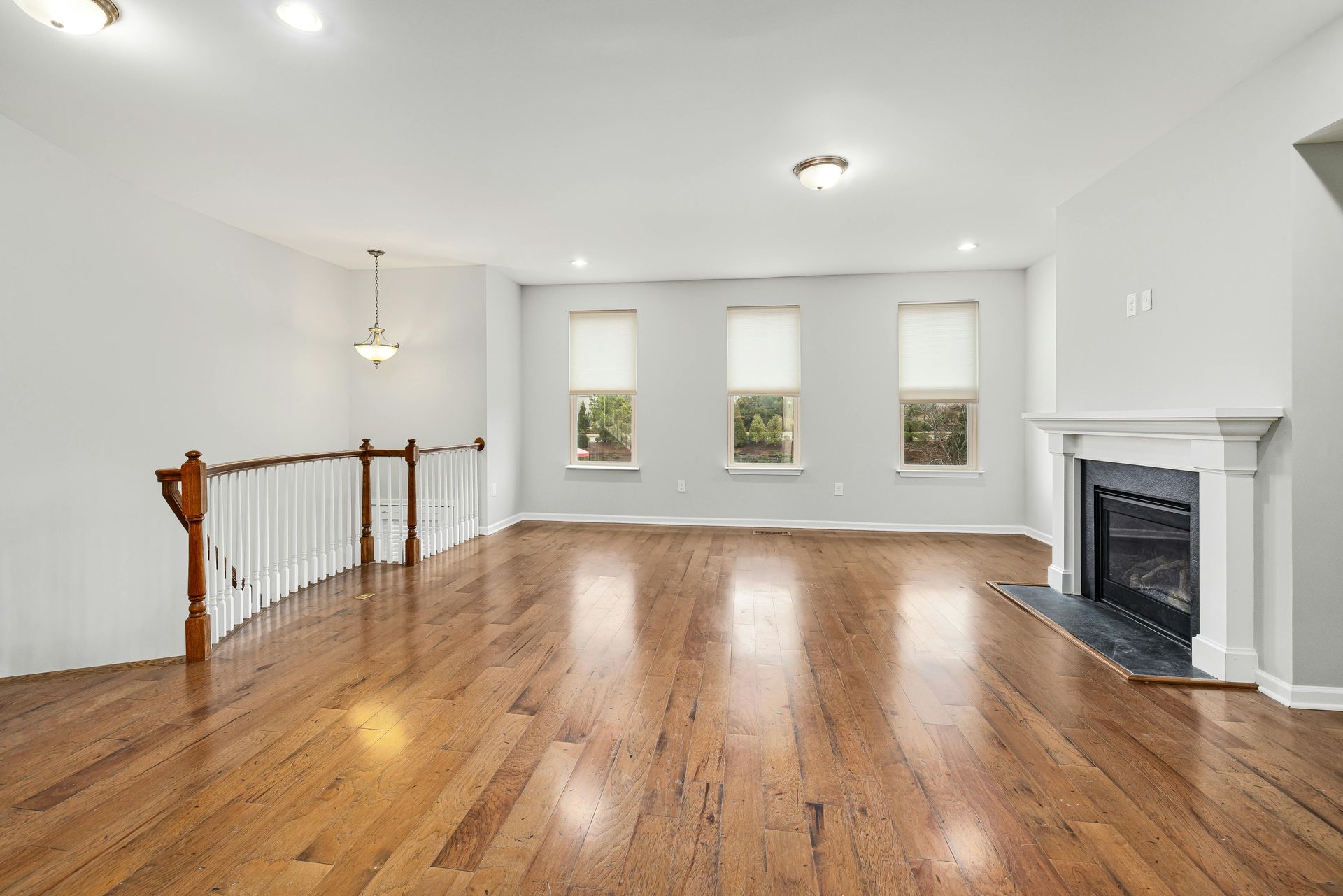 Empty living room with hardwood floors, three windows, a staircase, and a fireplace