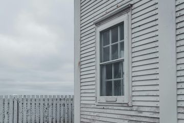 White house exterior with a window and weathered siding beside a white picket fence under a cloudy sky