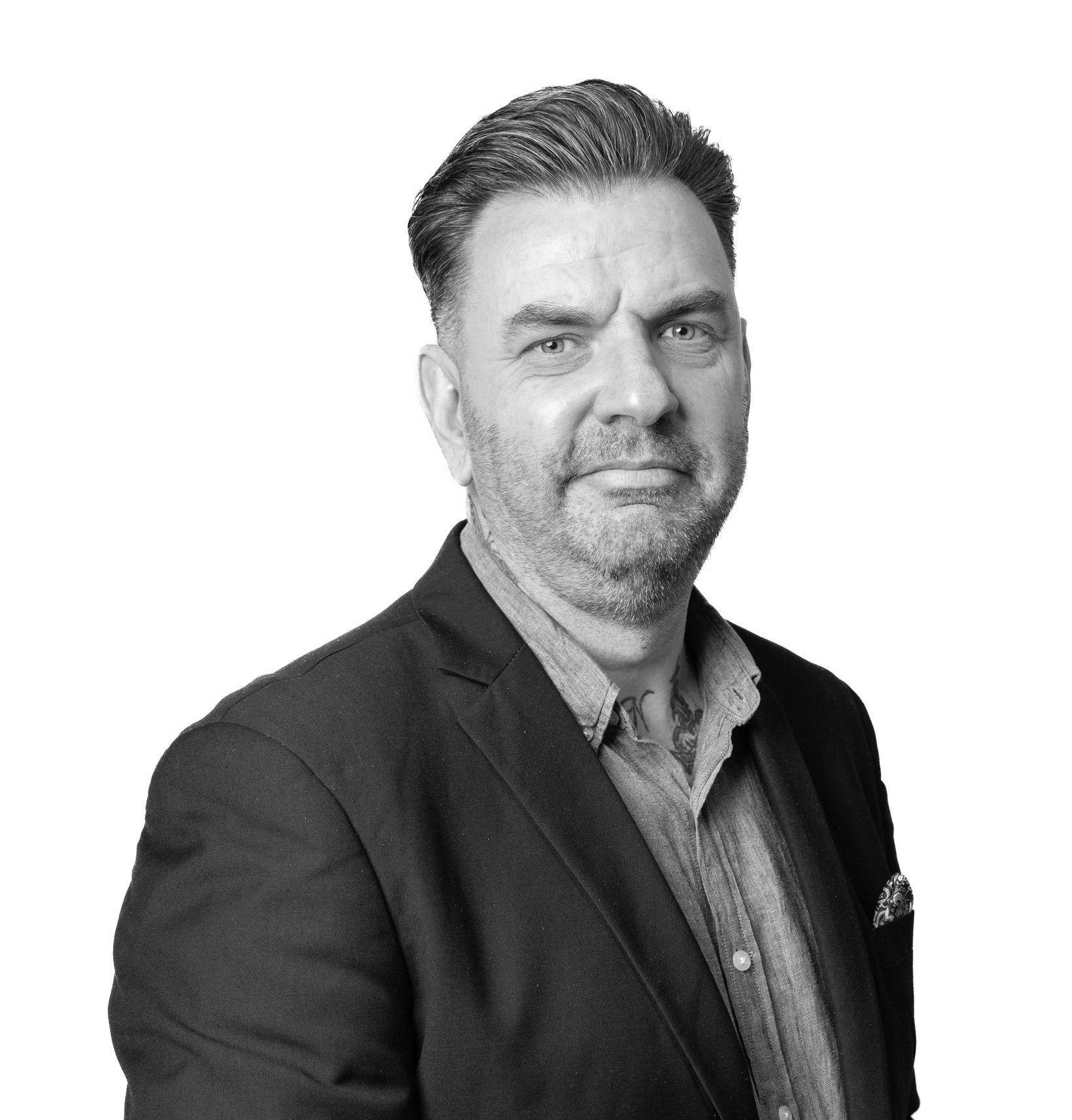 A man with a beard is wearing a suit and standing in front of a white background.