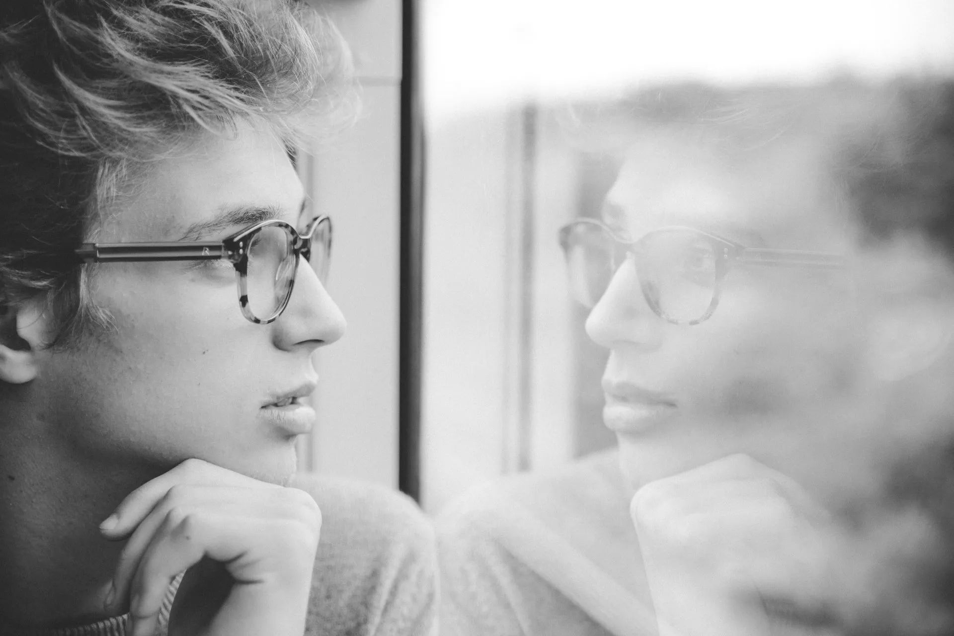 A black and white photo of a young man wearing glasses looking out a window.