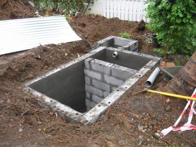 Partially built concrete utility pit in a dirt yard with pipes and a brick-lined wall