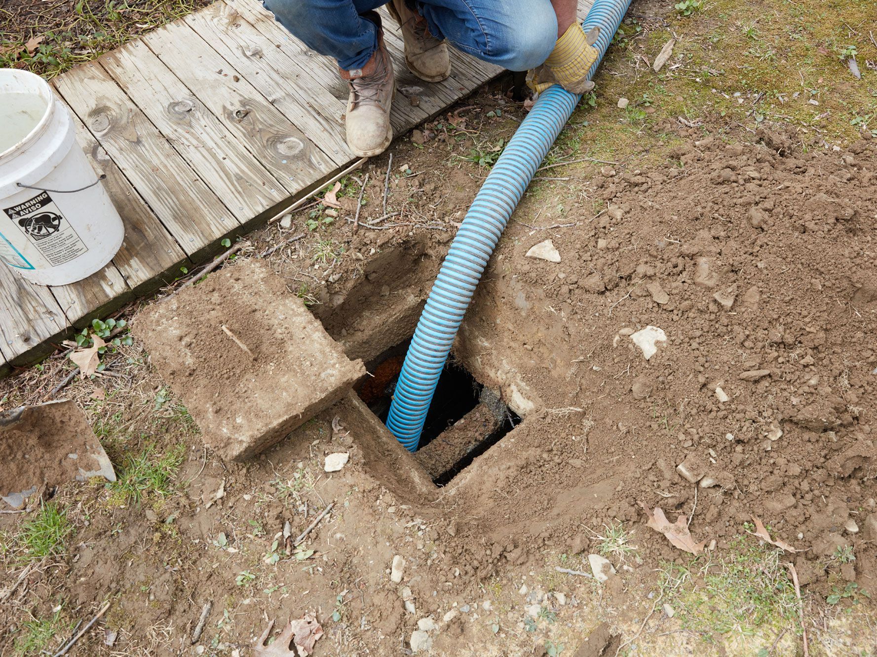 Person feeding a blue corrugated hose into a hole beside a wooden deck in a dirt yard