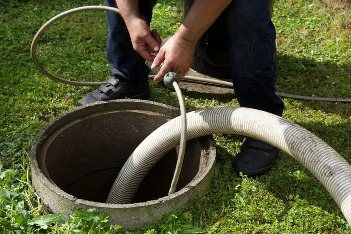 Person lowering a white hose into an open manhole in a grassy yard