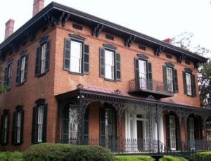 Historic two-story brick house with black shutters and a covered porch surrounded by greenery
