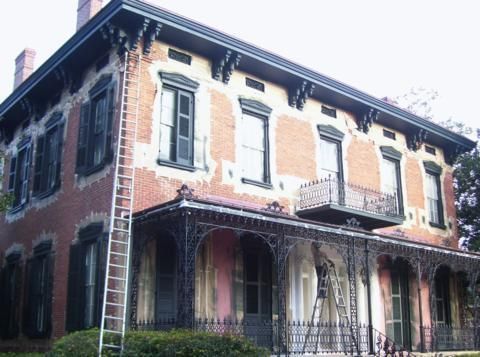 Old brick house with white trim, dark shutters, wrought-iron balconies, and a covered porch