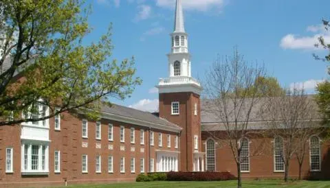 Brick campus building with white steeple and green lawn under a blue sky
