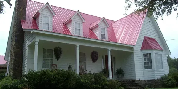 White house with a pink metal roof, front porch, and red dormer windows surrounded by greenery