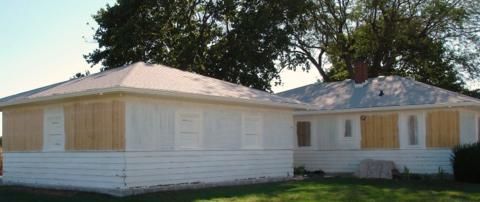 White single-story house with boarded-up windows and a damaged roof in a grassy yard