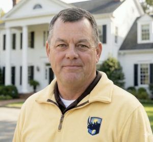 Man standing in front of a white house, wearing a pale yellow quarter-zip sweater.