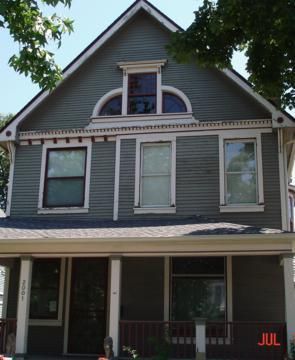 Gray two-story house with white trim, front porch, and arched attic window under trees.