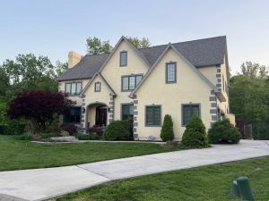 Large beige house with gray roof, arched entryway, and landscaped front yard beside a driveway