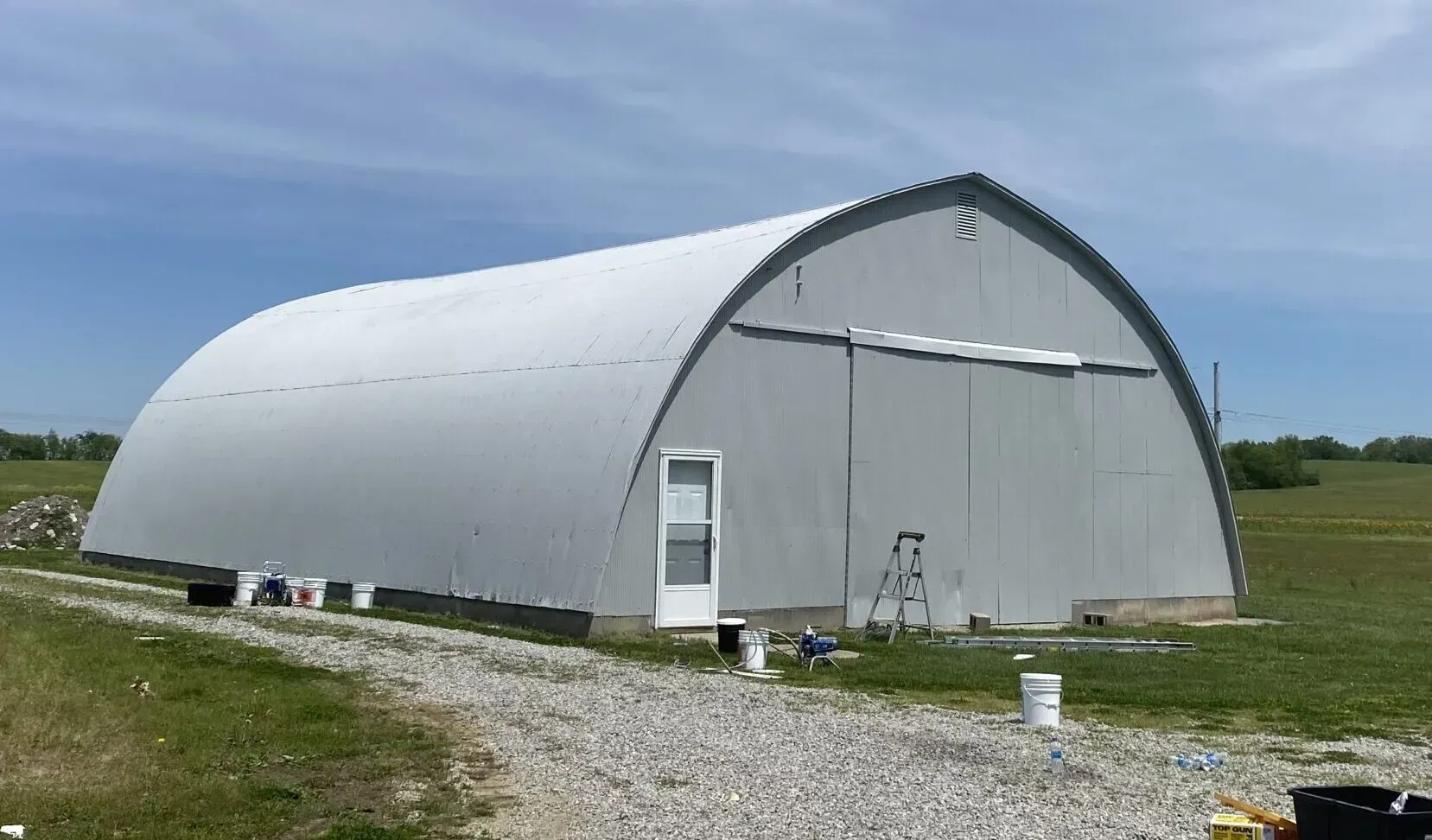 Large gray arched metal building on a gravel path in a grassy field under a blue sky