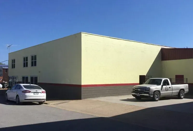 Small beige commercial building with red trim, a silver car parked beside it, and a pickup truck in front under a clear sky.