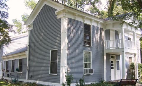 Blue-gray two-story house with white trim, a porch, and a small balcony surrounded by trees