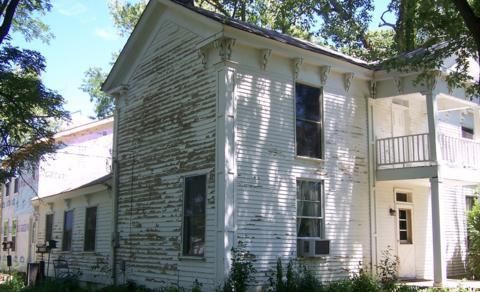 White two-story house with weathered siding, porch, and trees in the yard