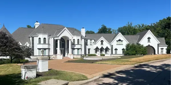 Large white mansion with columns and arched windows on a sunny street.