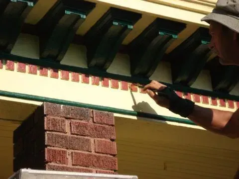 Person painting trim on a house exterior with a brush under the roof eaves.
