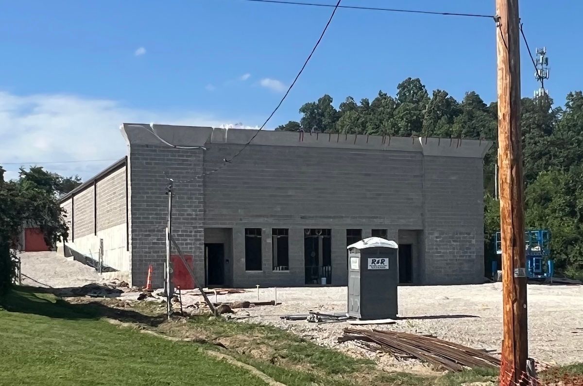 Gray commercial building under construction with gravel lot, utility pole, and portable toilet under a blue sky