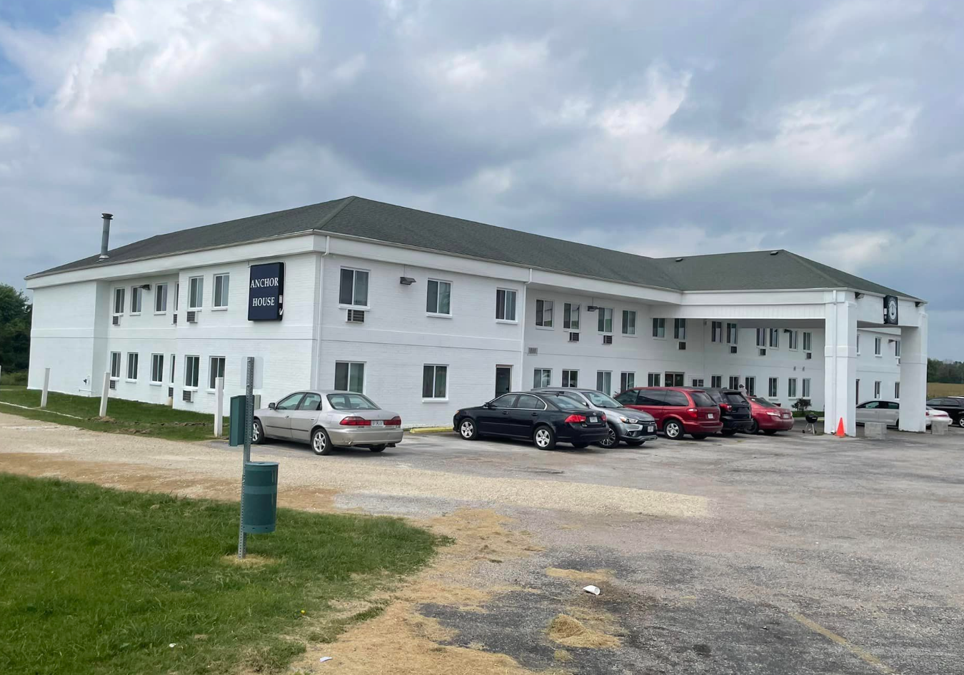 White two-story building with parked cars on a gravel lot under a cloudy sky.