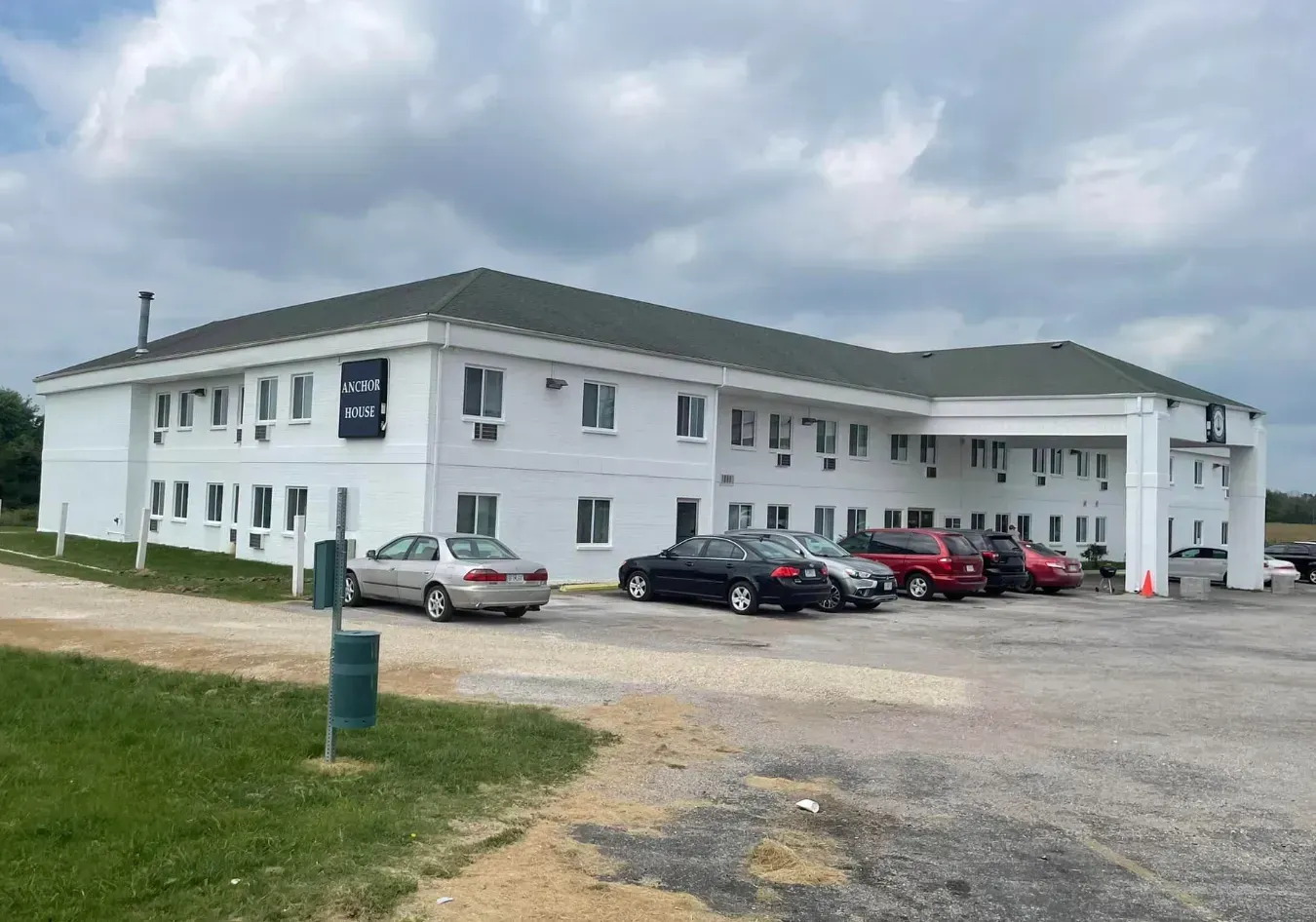 Two-story white building with green roof and parked cars on a gravel lot under cloudy sky