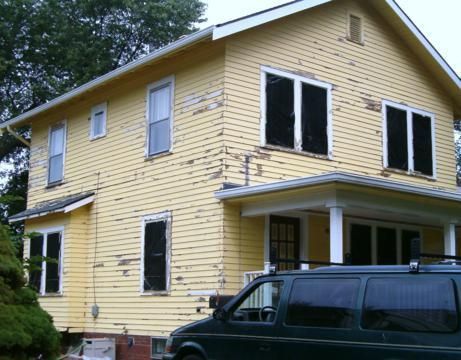 Yellow two-story house with white trim, damaged windows, and a black van parked in front