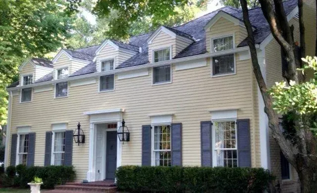 Beige two-story house with white trim, blue shutters, and dormer windows surrounded by trees.