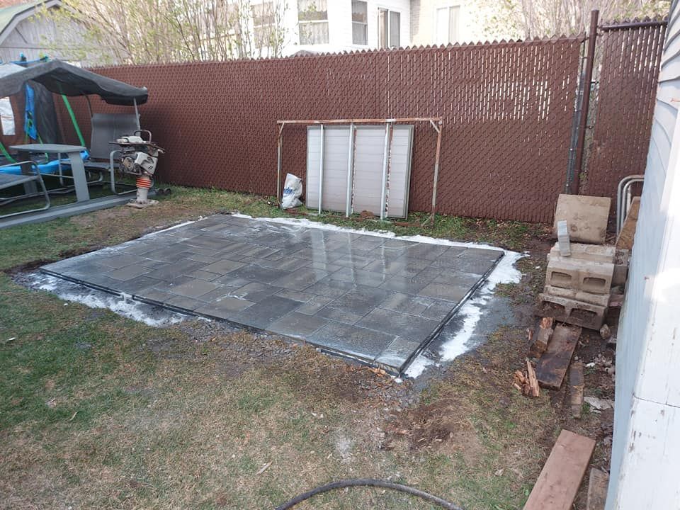 Une terrasse carrée et plate, pavée de dalles sombres et entourée de terre et d'herbe, se trouve dans un jardin. Une clôture brune est visible en arrière-plan.