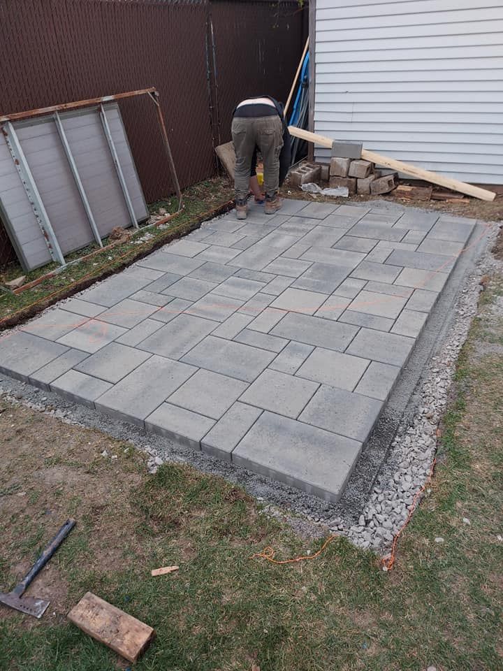 Une personne installe des pavés gris dans un jardin. La terrasse est entourée de gravier et de gazon.