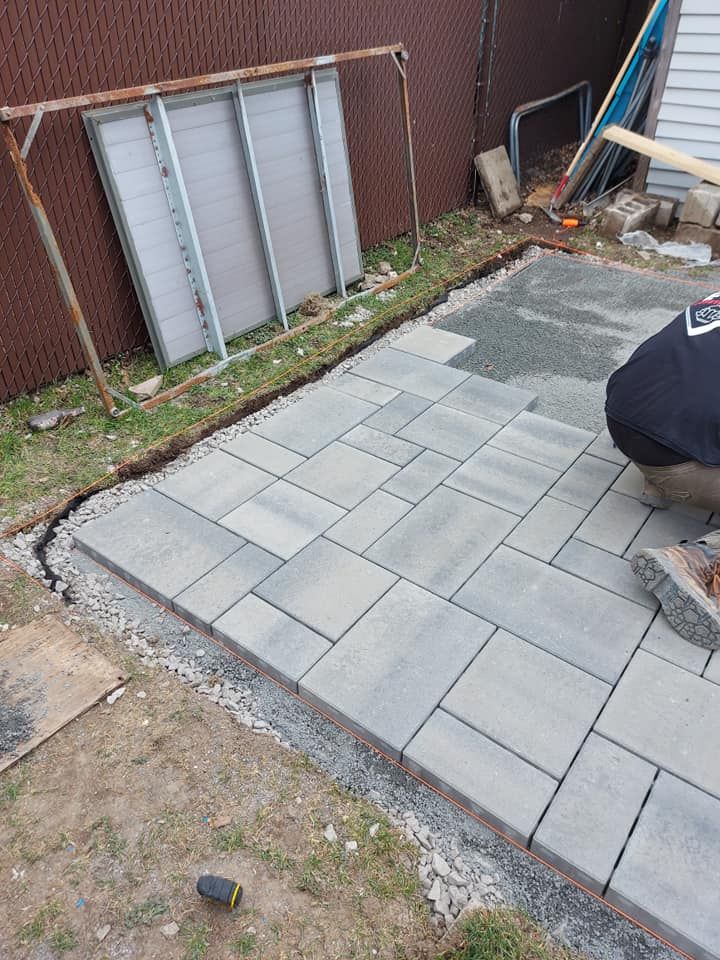Une personne pose des pavés sur une terrasse bordée de gravier. Une clôture brune et des matériaux de construction sont visibles en arrière-plan.