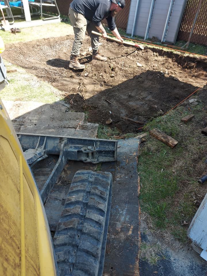 Une personne creuse un terrain vague en vue de travaux de construction. Une petite pelleteuse est visible au premier plan.