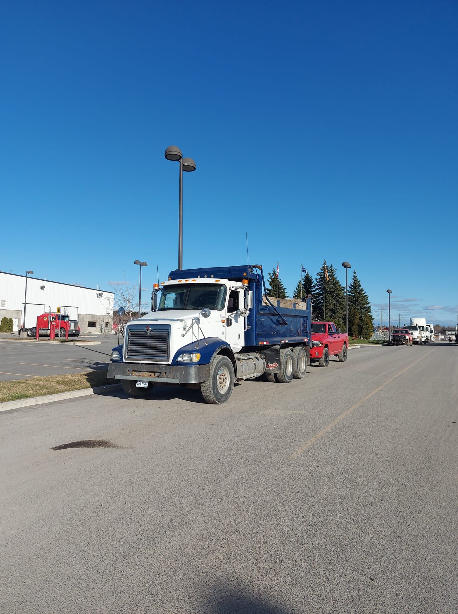 Un camion-benne blanc et bleu est stationné sur l'asphalte sous un ciel bleu azur. Une camionnette rouge y est attelée.