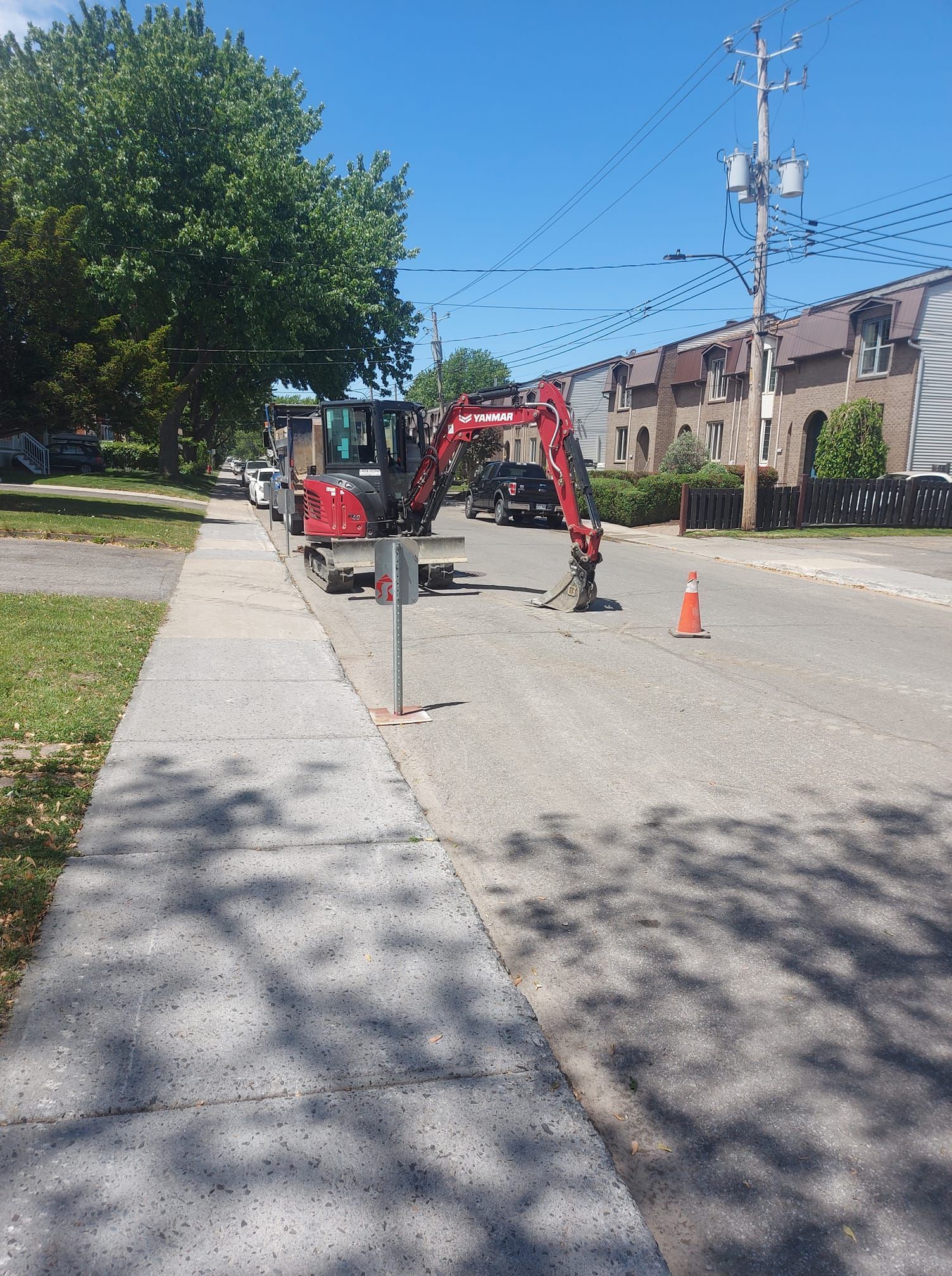 Chantier : petite pelleteuse sur une rue, le long d’un trottoir. Cône orange et poteau électrique visibles. Journée ensoleillée.