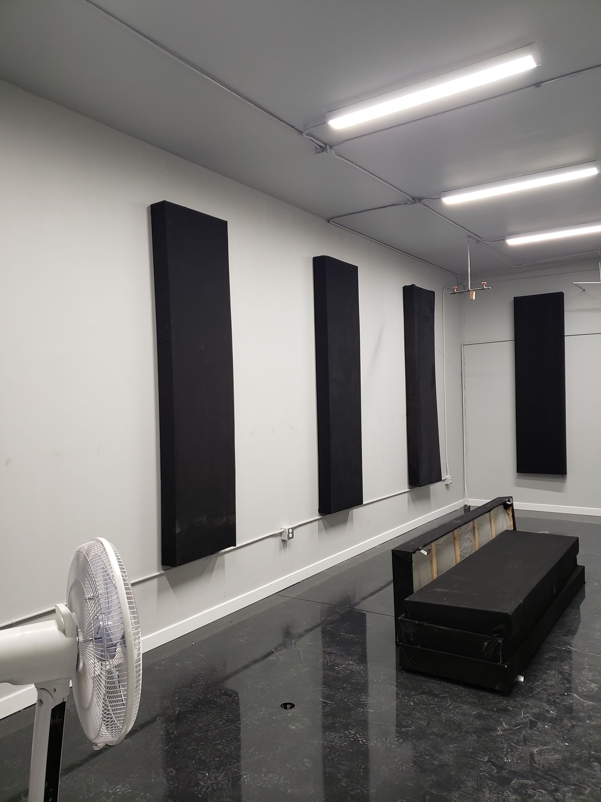Pièce blanche avec panneaux d'insonorisation au mur et tapis noirs empilés au sol, ventilateur au premier plan.
