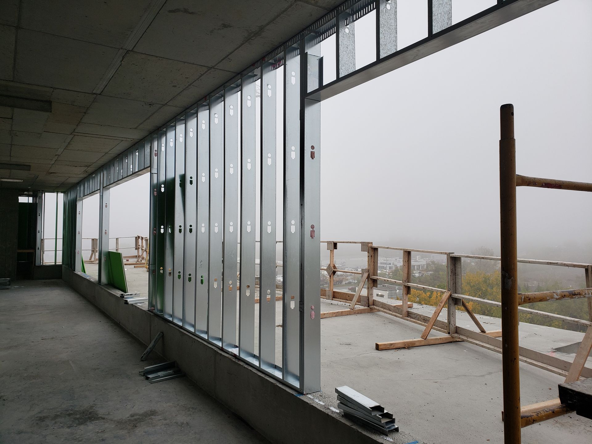 Vue intérieure d'un bâtiment en construction, montrant la structure métallique d'une grande fenêtre donnant sur une zone extérieure brumeuse.