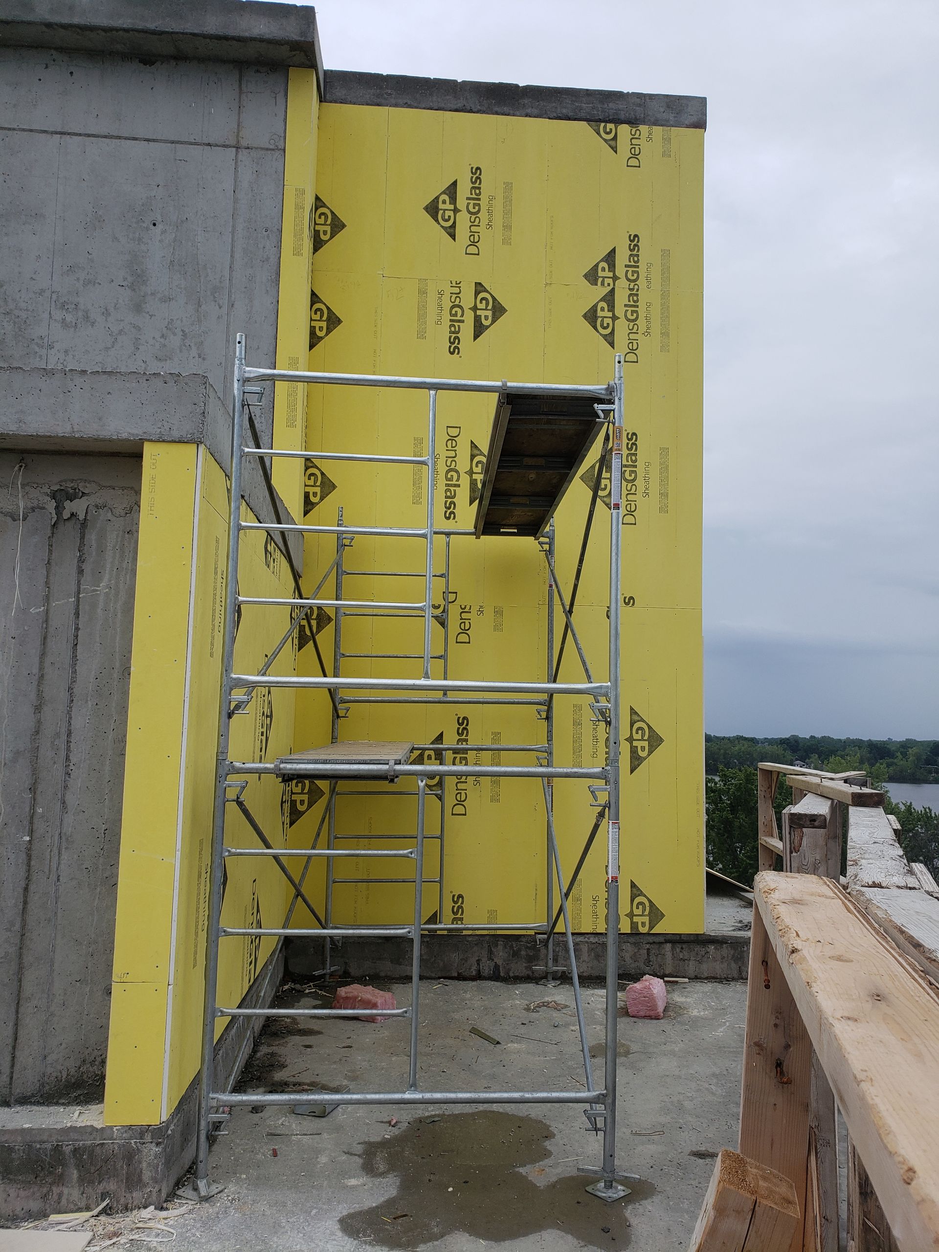 Échafaudage à côté d'un bâtiment en béton en construction, avec une barrière anti-intempéries jaune visible.