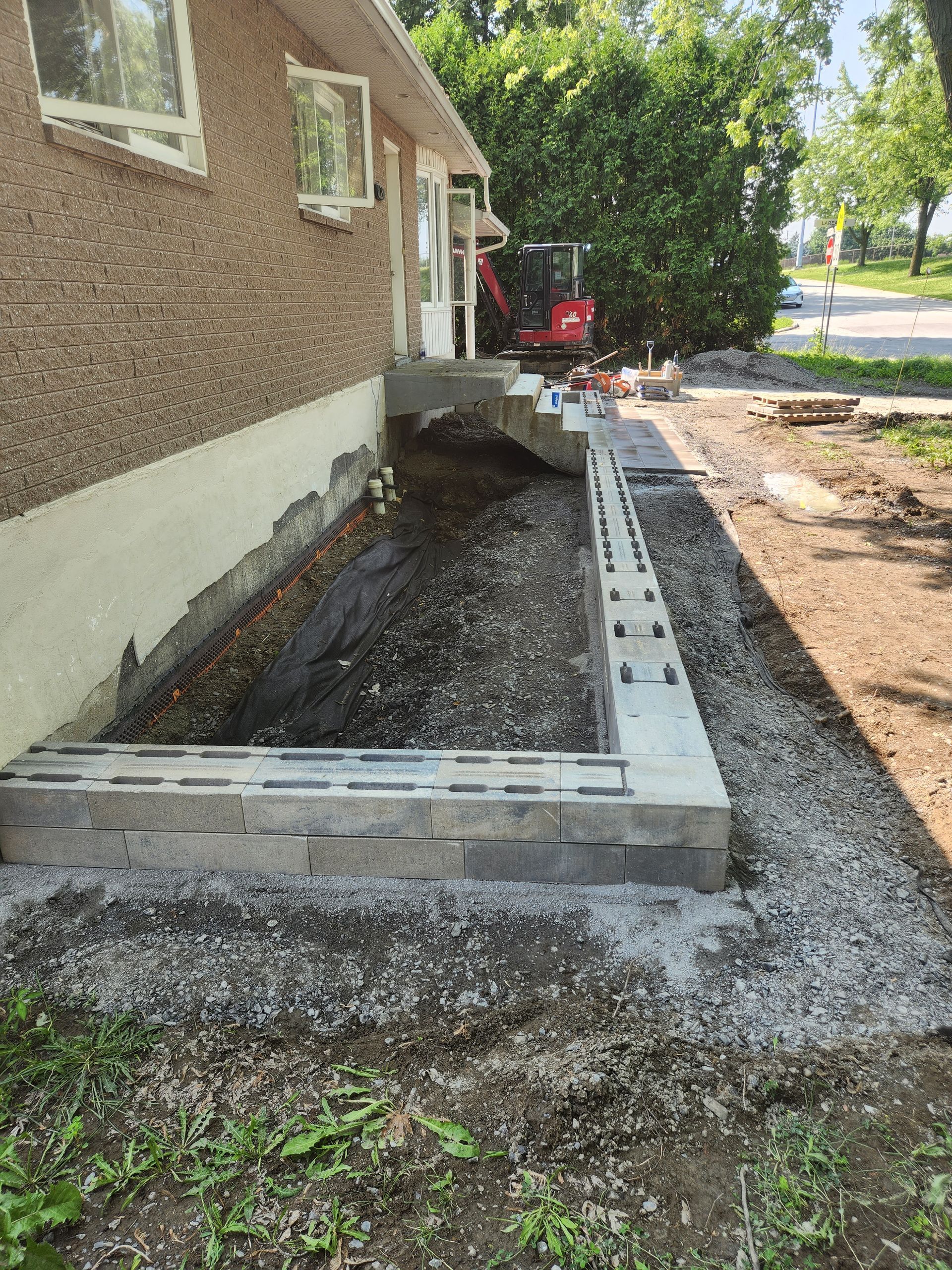 Une maison en construction. Un nouveau mur de soutènement et des marches sont en construction à côté de la maison, avec une mini-pelle en arrière-plan.