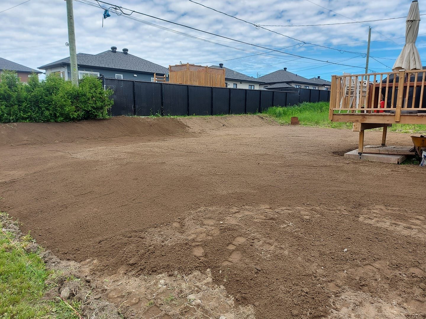Un jardin avec de la terre fraîche. Une terrasse en bois se trouve à droite, une clôture noire au fond, et des maisons sont visibles.