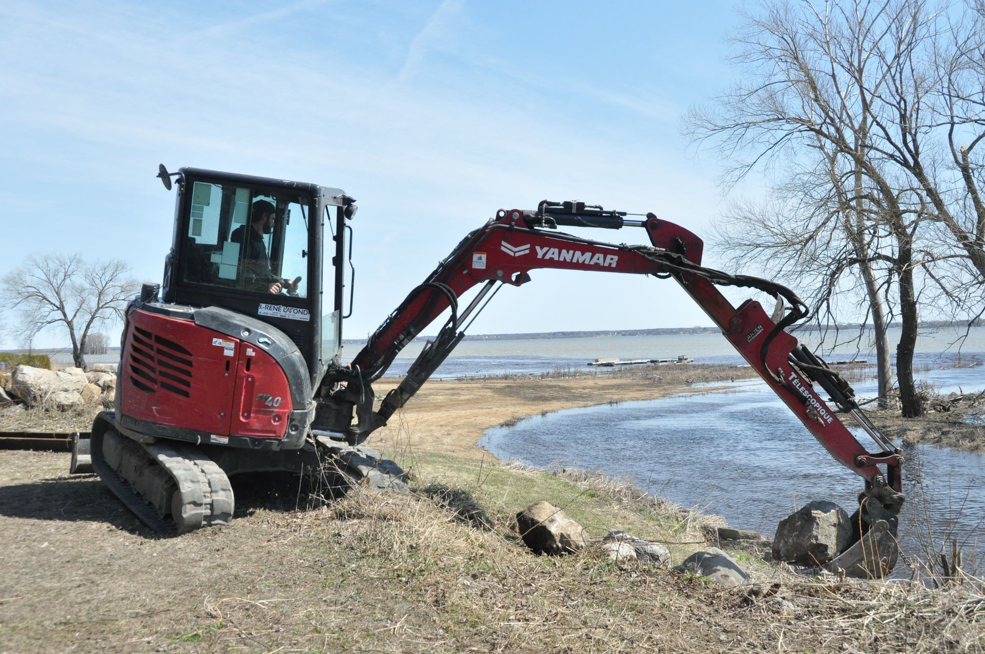 Une pelle mécanique Yanmar rouge, en train de creuser près de l'eau sur le rivage, avec des arbres et un ciel nuageux en arrière-plan.