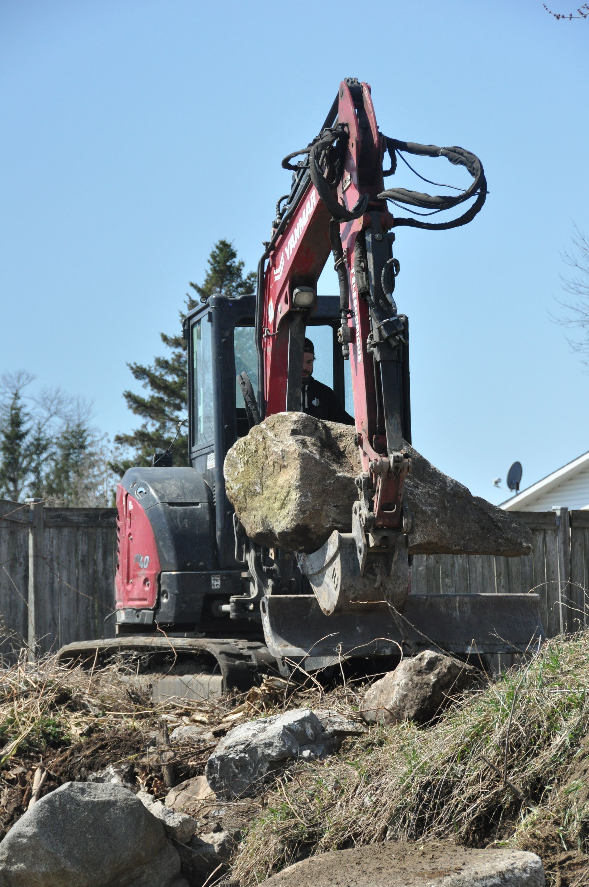 Une pelleteuse rouge soulève un gros rocher à l'extérieur, près d'une clôture en bois, par une journée ensoleillée.