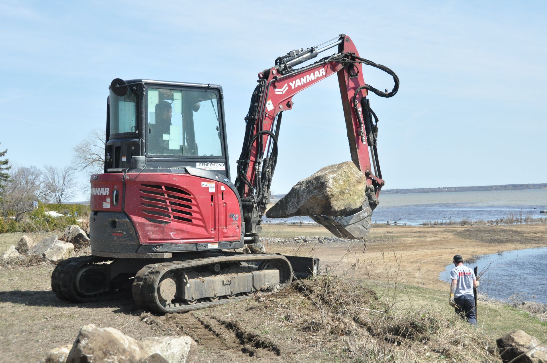 Une pelleteuse rouge soulève un gros rocher sur le littoral ; une personne observe à proximité.