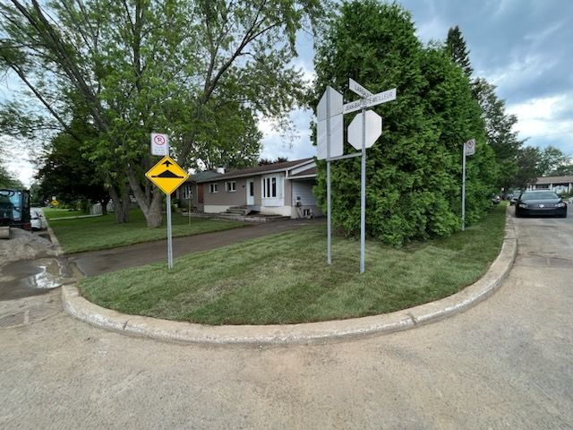 Bordure de trottoir à un carrefour résidentiel avec panneaux : ralentisseur, stop et noms de rue. Pelouse verte et maison grise.