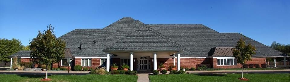 Single-story brick building with a gray shingled roof and white columns, surrounded by a lawn and trees.