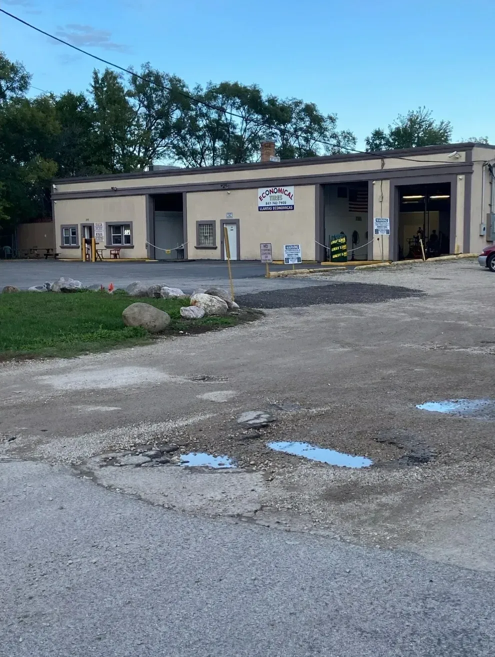 Exterior view of a beige building, possibly a fire station. Paved area in the foreground with puddles. Trees in the background.