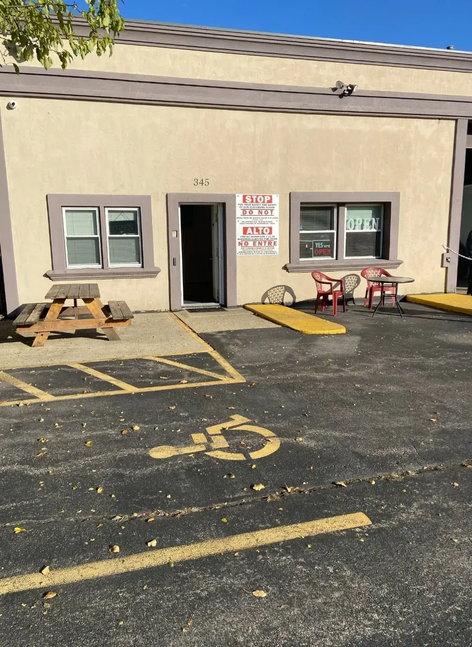 Exterior of a beige building with a dark open doorway, a wheelchair accessible ramp, and tables and chairs outside.