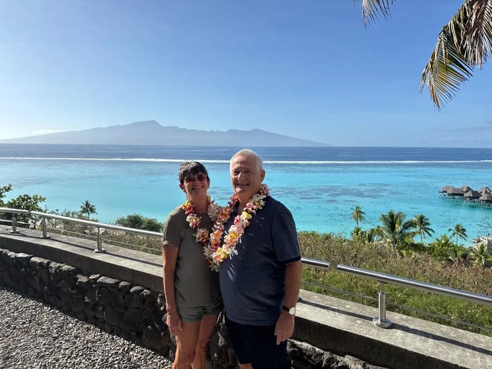 Two people wearing leis stand by a seaside overlook with ocean, mountain, and blue sky in the background