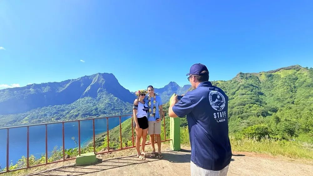 Two hikers pose on a mountain overlook with lake and green hills under a clear blue sky.