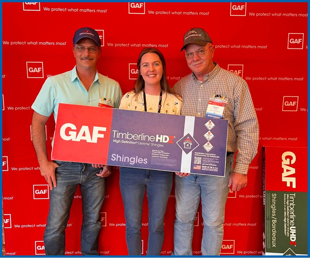 Three people hold a GAF Timberline HD Shingles check at a red GAF event backdrop.