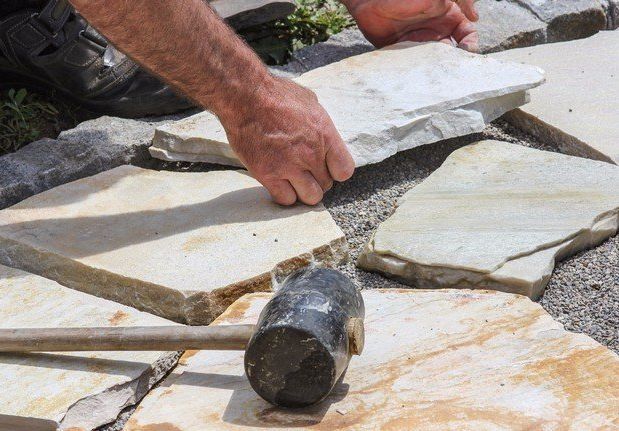 Hands laying irregular stone slabs with a mallet on a stone patio.